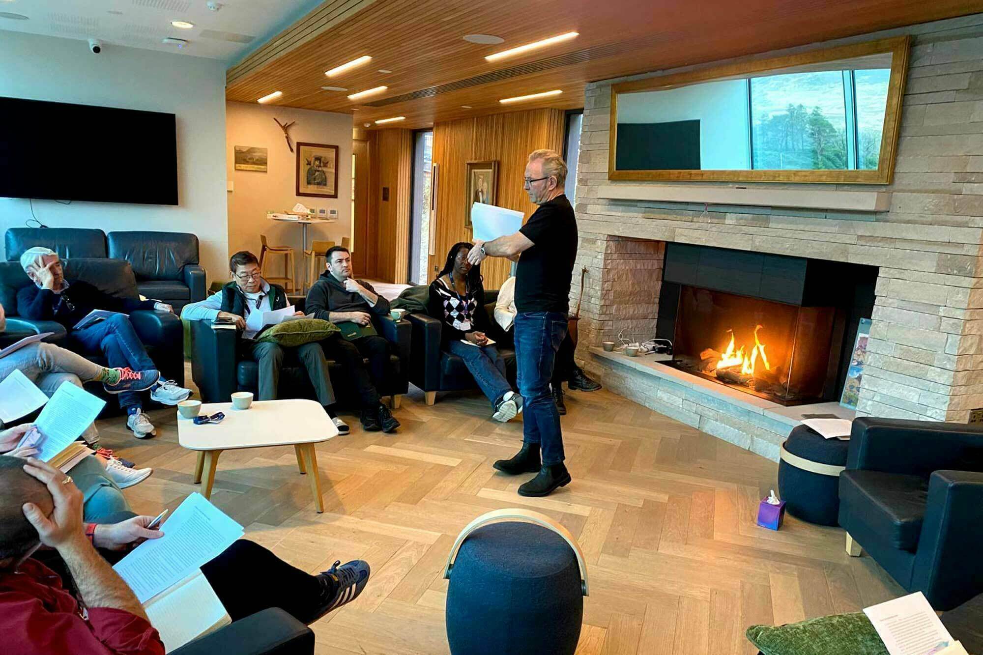 A man stands holding papers, addressing a small group seated in a lounge area. Participants sit in armchairs, some holding documents or books, engaged in the discussion. A fireplace with a burning fire adds warmth to the wood-floored room. A large mirror above the fireplace reflects the outside scenery.