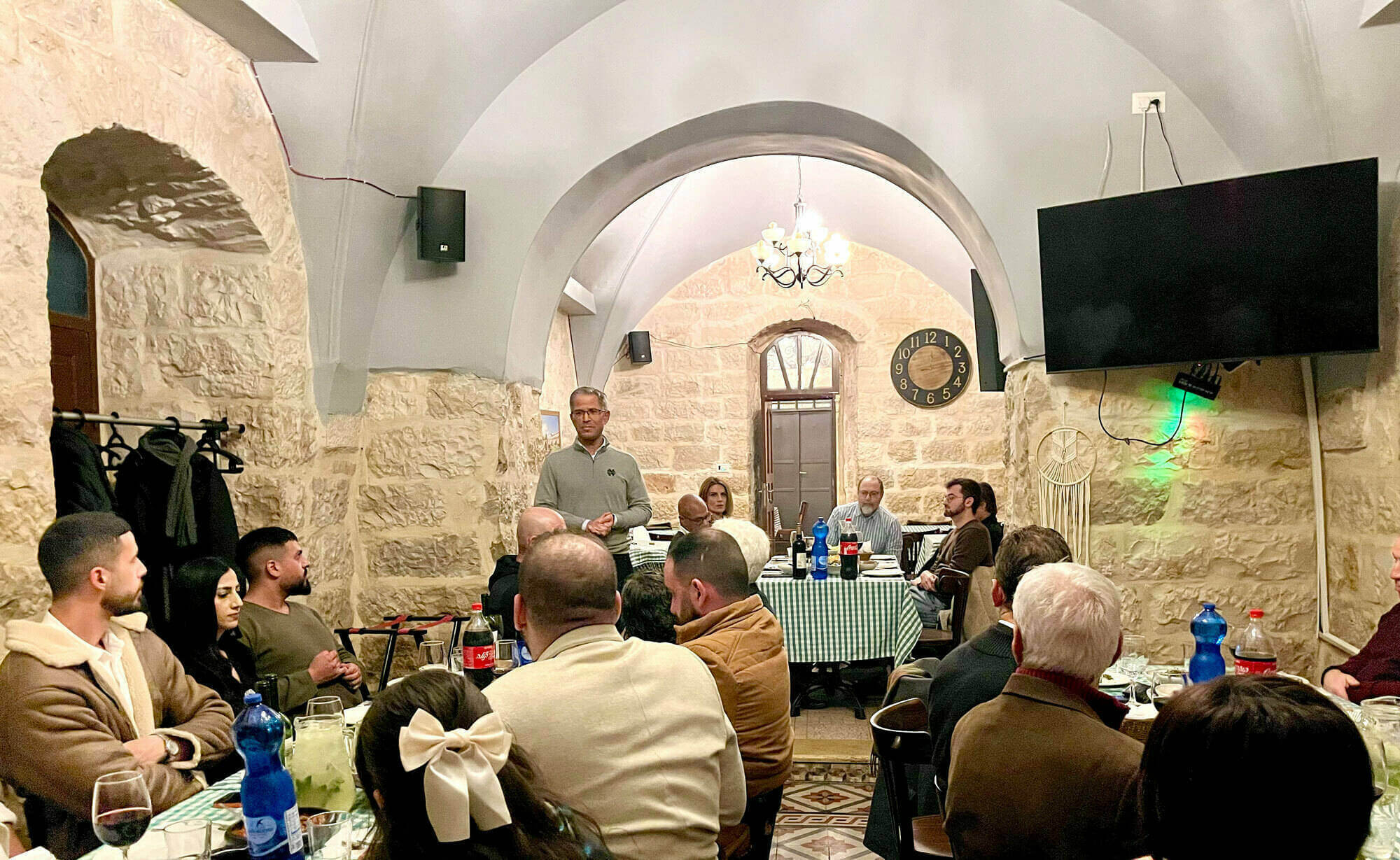 Daniel Schwake, executive director of ND Jerusalem stands speaking to staff members seated at tables in a stone-walled room with arched doorways and ceilings. A clock and a flat-screen TV are mounted on the walls.