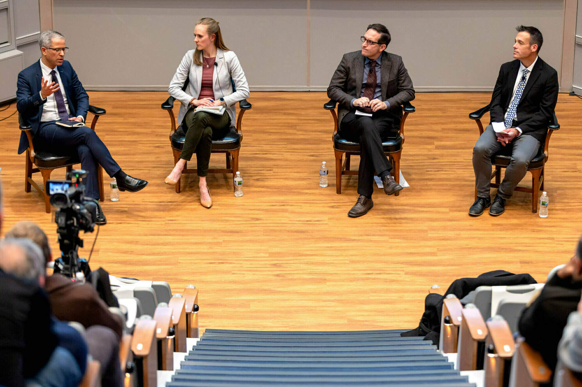 Four panelists sit in chairs on a stage, speaking to an audience in a lecture hall.  A video camera records the event.