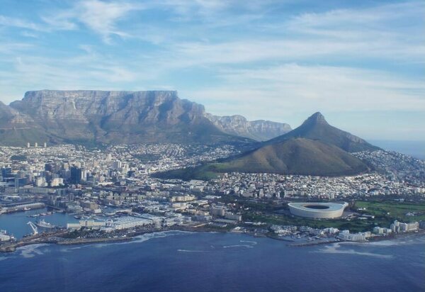 Aerial view of Cape Town, South Africa, with Table Mountain in the background, Lion's Head to the right, and the city sprawling towards the coastline.  The ocean is a deep blue, and a large stadium is visible near the shore.