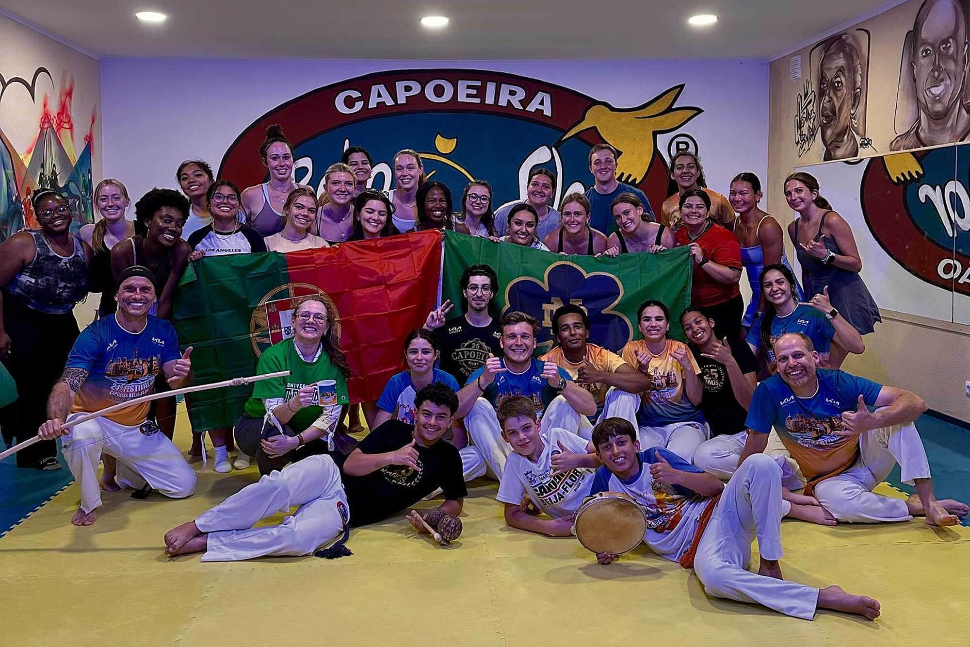 A large group of people pose for a photo in a Capoeira studio. They hold the Portuguese and Brazilian flags and give thumbs-up gestures. Some wear Capoeira uniforms and hold instruments. Murals decorate the walls.