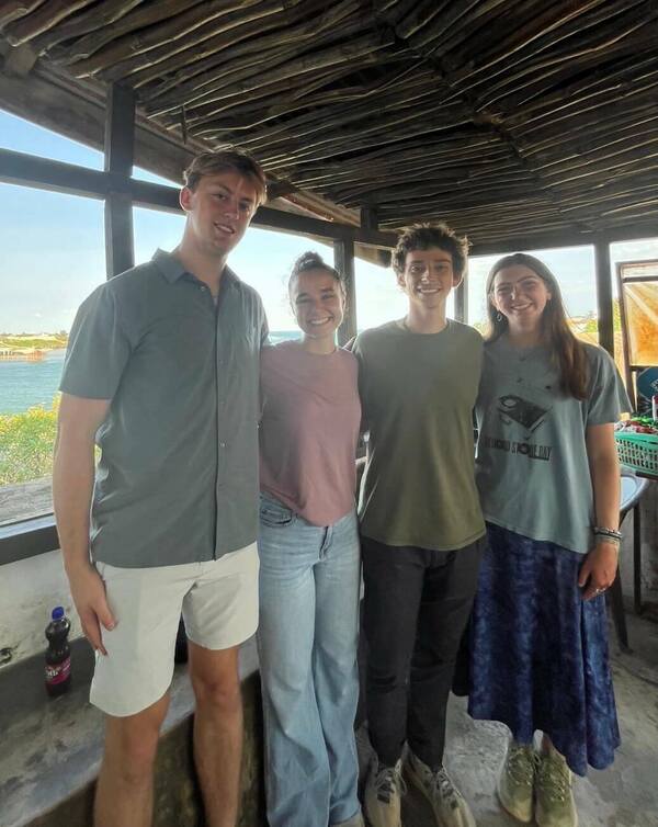 Four students smile for a photo inside a thatched-roof structure with a view of water in the background.