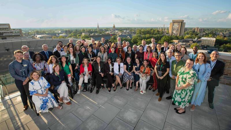 A large, diverse group of people smiles for a photo on a rooftop patio with Notre Dame Stadium, the Golden Dome, Basilica steeple, and Hesburgh Library visible in the sunny background.