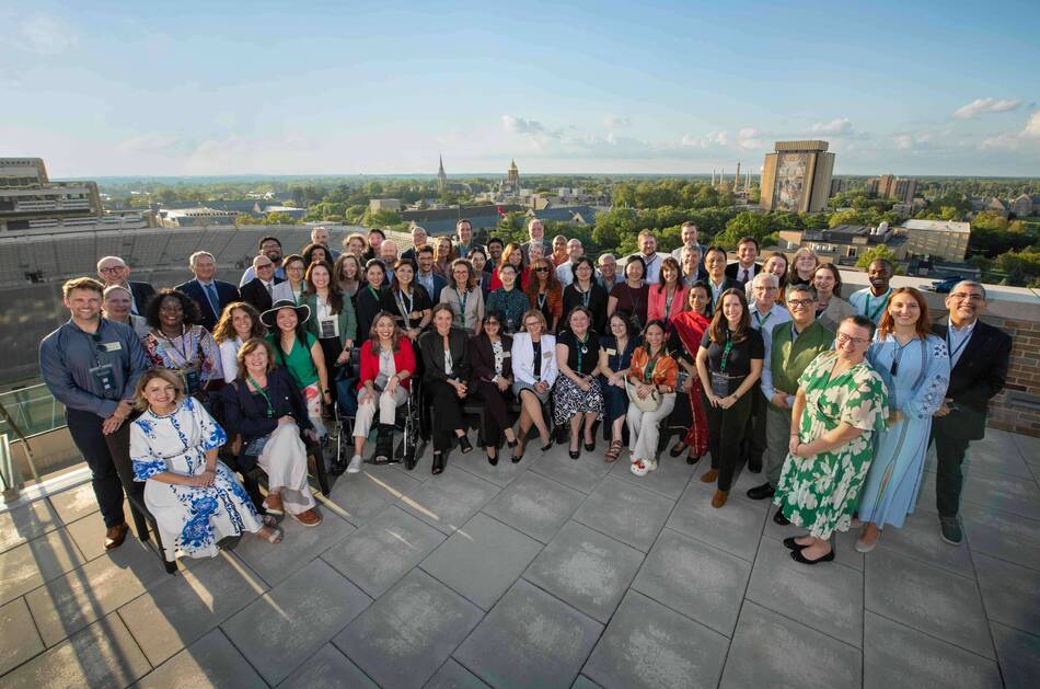 A large, diverse group of people smiles for a photo on a rooftop patio with Notre Dame Stadium, the Golden Dome, Basilica steeple, and Hesburgh Library visible in the sunny background.
