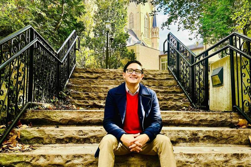 A smiling man with glasses, wearing a navy coat and red sweater, sits on rugged stone steps with black railings. The Basilica of the Sacred Heart rises behind autumn trees in the background.