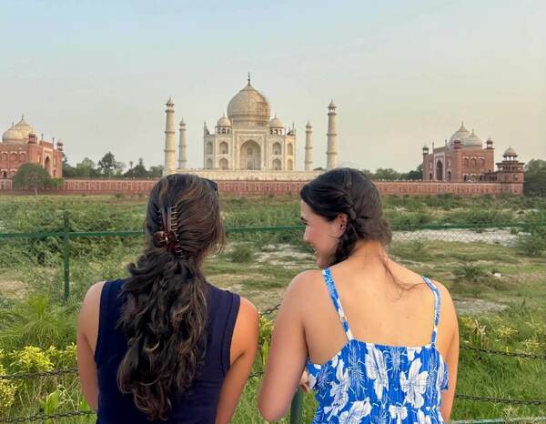 Two women from behind gaze at the white marble Taj Mahal, flanked by red sandstone buildings. The woman on the left wears a navy top; the right, a blue and white floral dress, smiles.