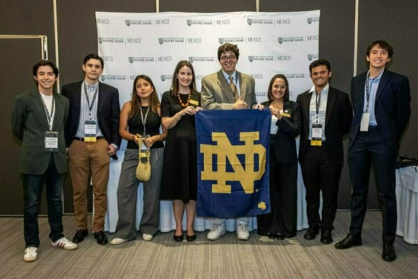 Eight smiling individuals, five men and three women, in business casual attire, proudly hold a large blue and gold Notre Dame flag before a repeating University of Notre Dame Mexico logo backdrop. One man gives a thumbs up.