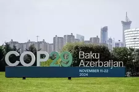 Large white and green COP29 sign on a dark blue base in a grassy field. It reads "COP29 Baku Azerbaijan November 11-22 2024". A city skyline with multiple buildings and green trees is visible in the background.