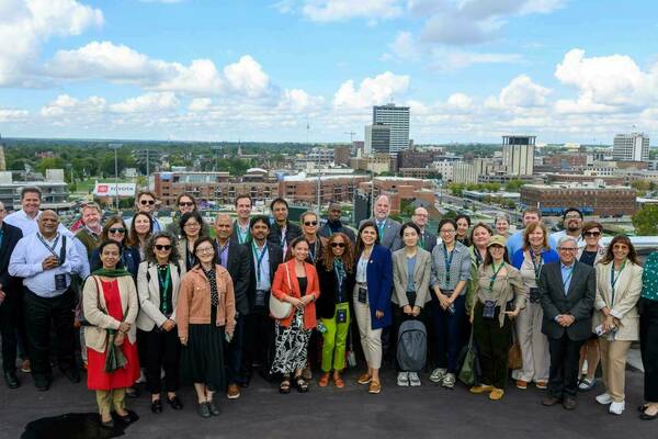 A large, diverse group of smiling people stand on a rooftop. They wear lanyards and overlook a city skyline with buildings, a church steeple, and a Toyota sign, under a partly cloudy sky.