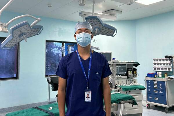 A young man in dark blue scrubs, a light blue surgical cap, and mask stands, hands in pockets, looking at the camera in an operating room. Surgical lights, a green-draped table, and monitors surround him.