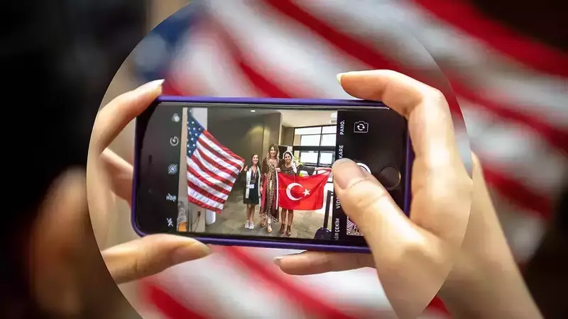 A person's hands hold a smartphone, displaying a photo of three smiling women. One woman holds an American flag, another a red Turkish flag with a white crescent moon and star. The overall image background is a blurred American flag.