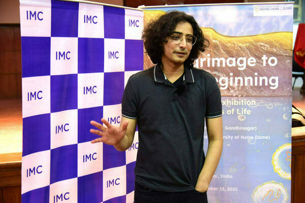 A young man with dark, curly hair and round glasses, wearing a charcoal grey polo, gestures. He stands between a blue and white "IMC" banner and a "University of Notre Dame Global" exhibition banner.