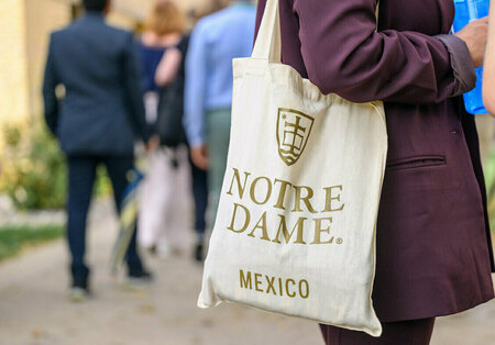 A person in a burgundy blazer carries a natural canvas tote bag featuring the gold Notre Dame logo and "NOTRE DAME MEXICO" text, walking past blurred people outdoors.
