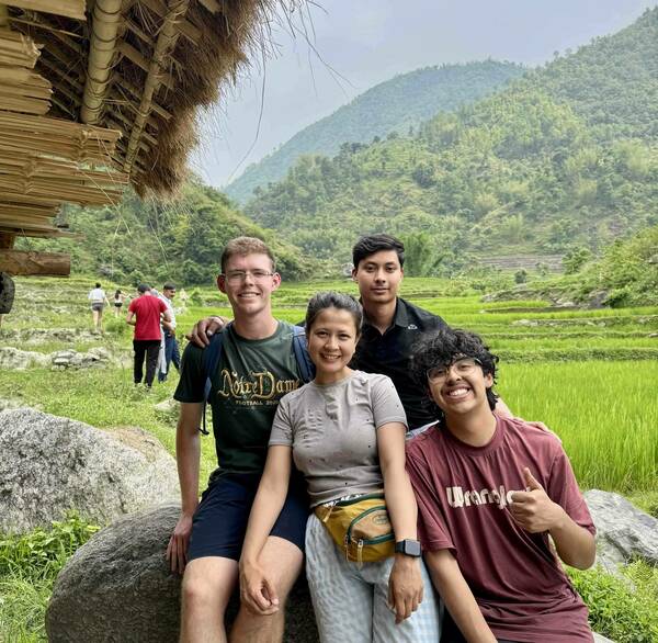 Four smiling young people sit on rocks. One wears a green "Notre Dame Football 2025" shirt. Behind them are vibrant green rice paddies, lush mountains, and a cloudy sky. The person on the right, wearing a maroon shirt, gives a thumbs-up. A thatched roof is visible on the left.