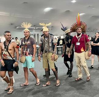 Five indigenous men, adorned in diverse traditional attire including feather headdresses, beaded necklaces, and body paint, walk through an indoor hall. The man in red smiles, wearing a vibrant headdress and Brazilian flag patch.