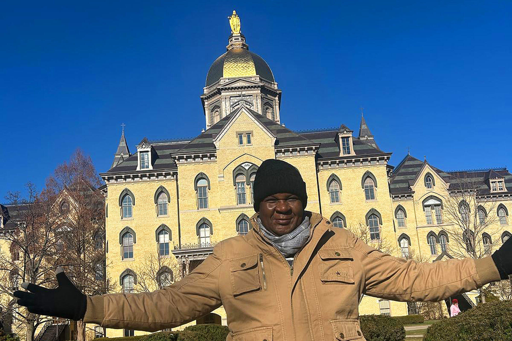 A smiling man in a black beanie, grey scarf, and tan jacket with outstretched arms, stands before Notre Dame's iconic Main Building, featuring its yellow brick and Golden Dome, against a bright blue sky.