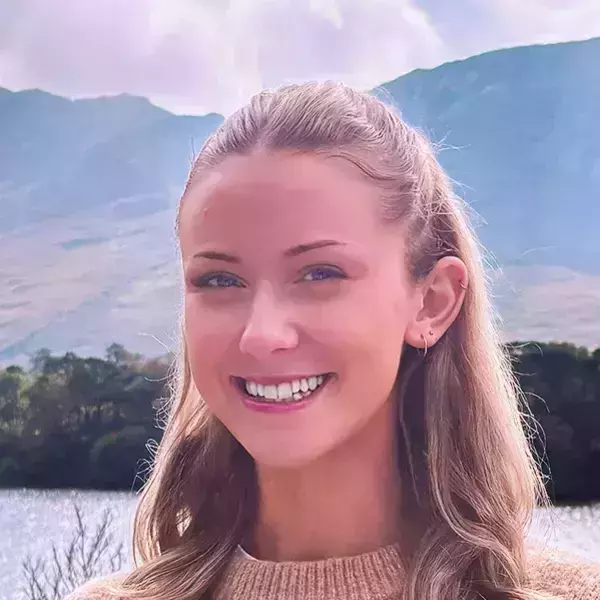 Lily Mansfield. Smiling young woman with blonde hair and a brown sweater, framed by a lake and blue mountains under a cloudy sky.