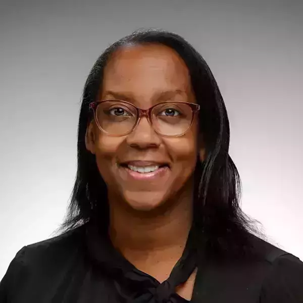 Cynthia Reeves. A smiling woman with dark skin and long dark hair wears reddish-brown glasses and a black blouse with a tied neckline.