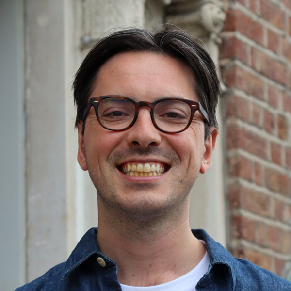 Patrick Schmitz. A smiling man with dark hair and brown glasses wears a blue shirt over a white t-shirt. He stands against a background of light stone carving and red brick.