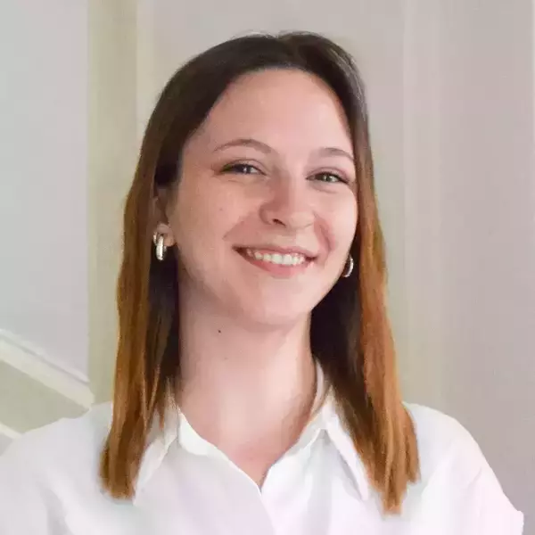 Aurora Nolfo. Young woman with reddish-brown hair, light skin, and freckles smiles broadly, wearing a white collared shirt and silver earrings.