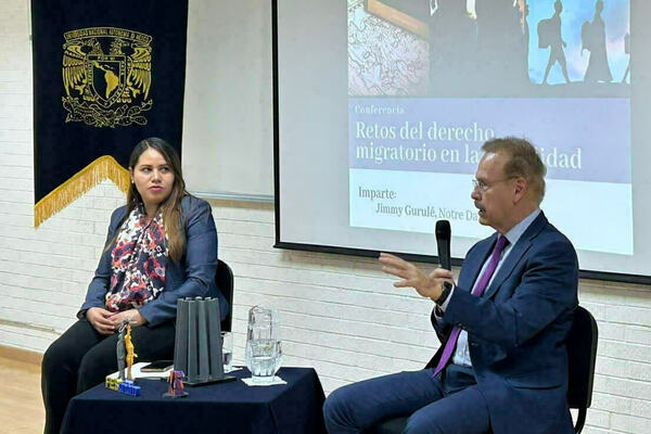 Jimmy Gurulé speaks in blue suit/purple tie, holding a mic, as woman listens. Screen shows migration law topic.