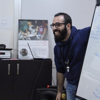A man with a dark beard, black glasses, and a blue hoodie laughs heartily while looking at a whiteboard.