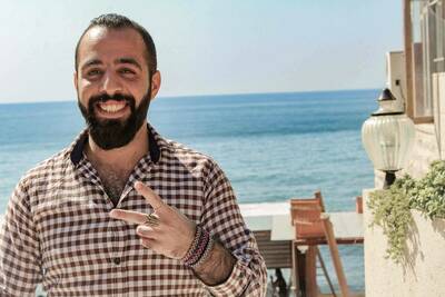 Smiling man with a dark beard in a brown checkered shirt making a peace sign, with the ocean and blue sky behind him.