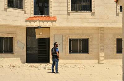 Man in dark vest and jeans looks right at a light-colored building with a broken upper window and ground debris.