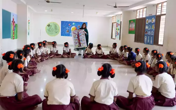A teacher in green presents a tree poster to young girls in cream/maroon uniforms with orange hair ties, sitting in a circle.