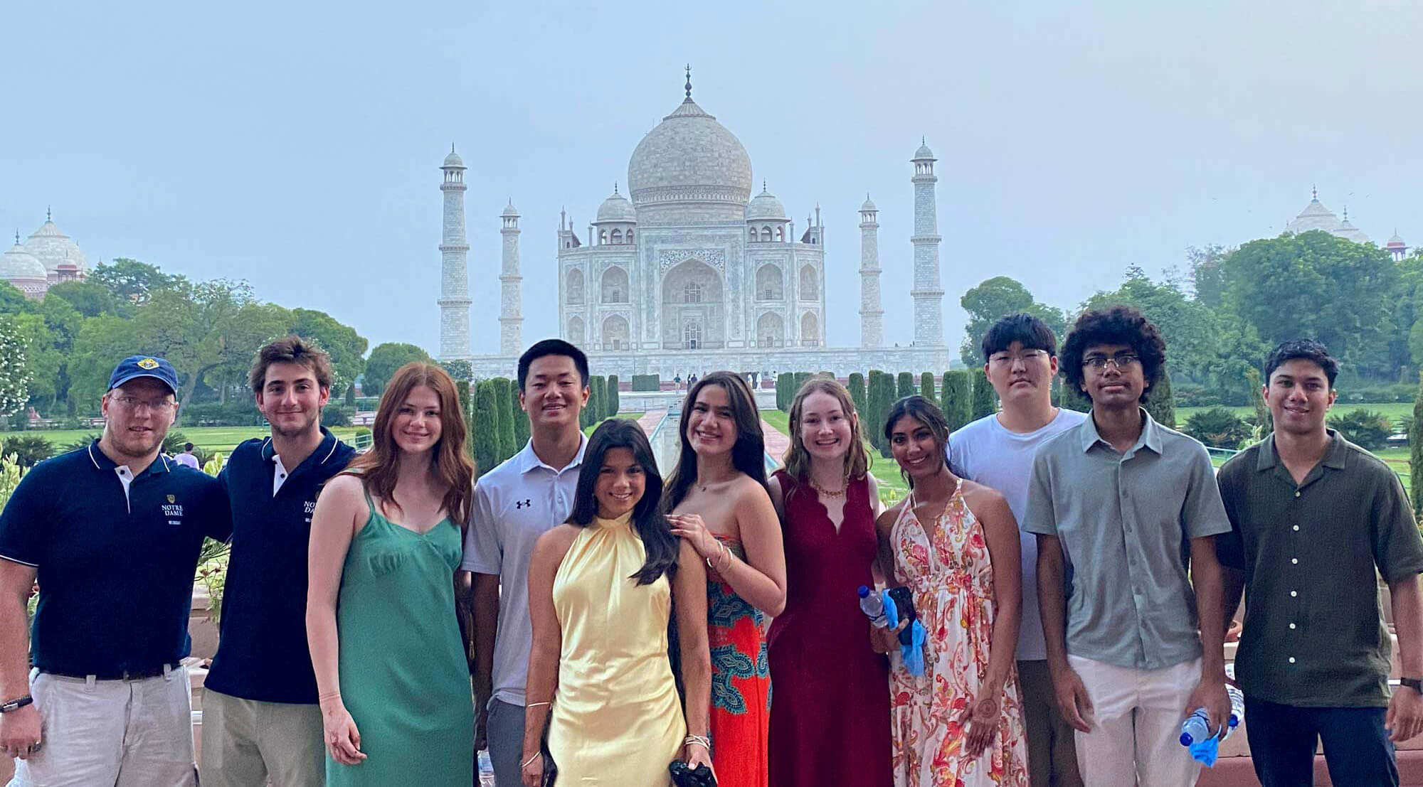 Ten smiling individuals, some in Notre Dame attire, pose before the white marble Taj Mahal's dome and minarets.