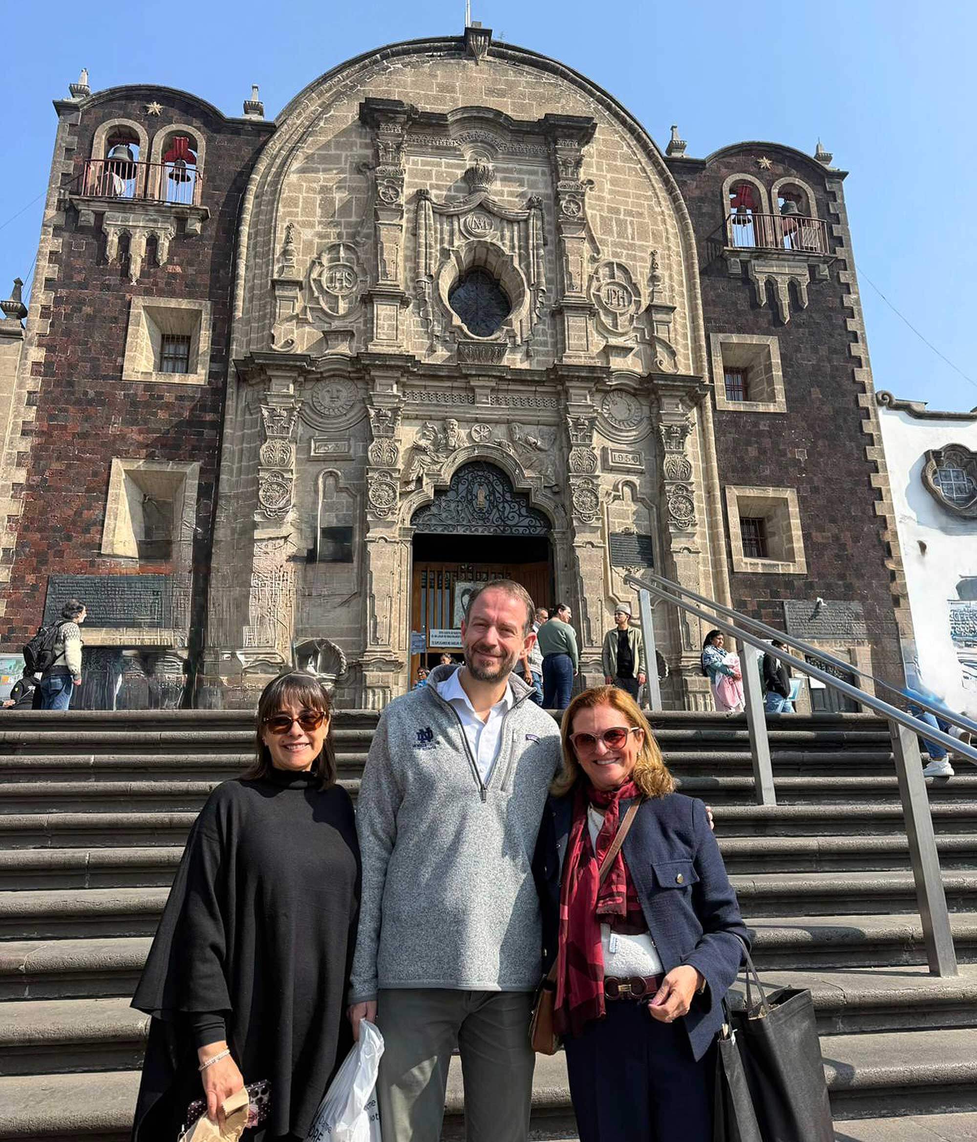 Three smiling people, including a man in a grey Notre Dame sweatshirt, stand on stone steps in front of an ornate church.