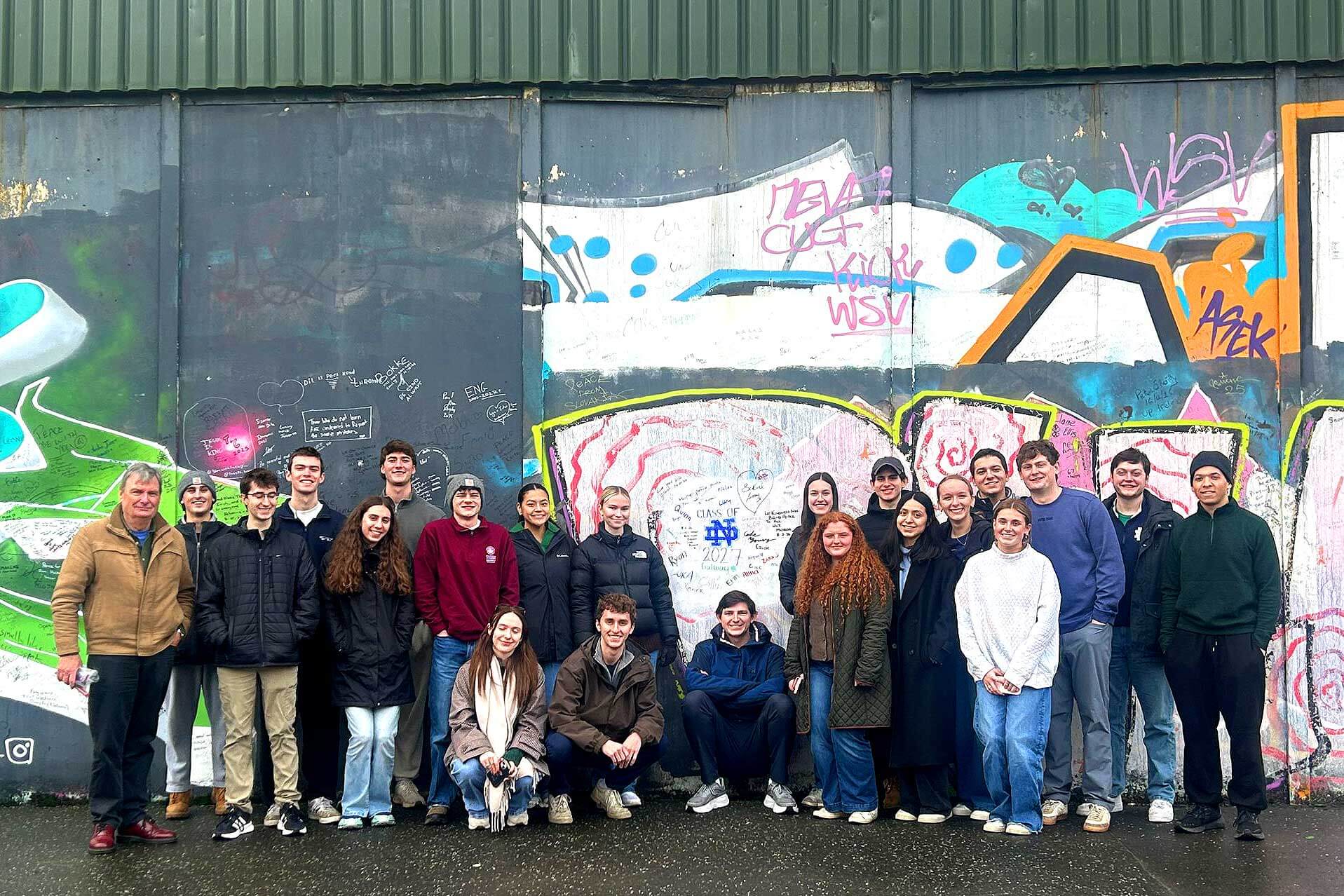 A group of Notre Dame students and a man pose, smiling, in front of a graffiti-covered wall with "Class of ND 2027" written on it.