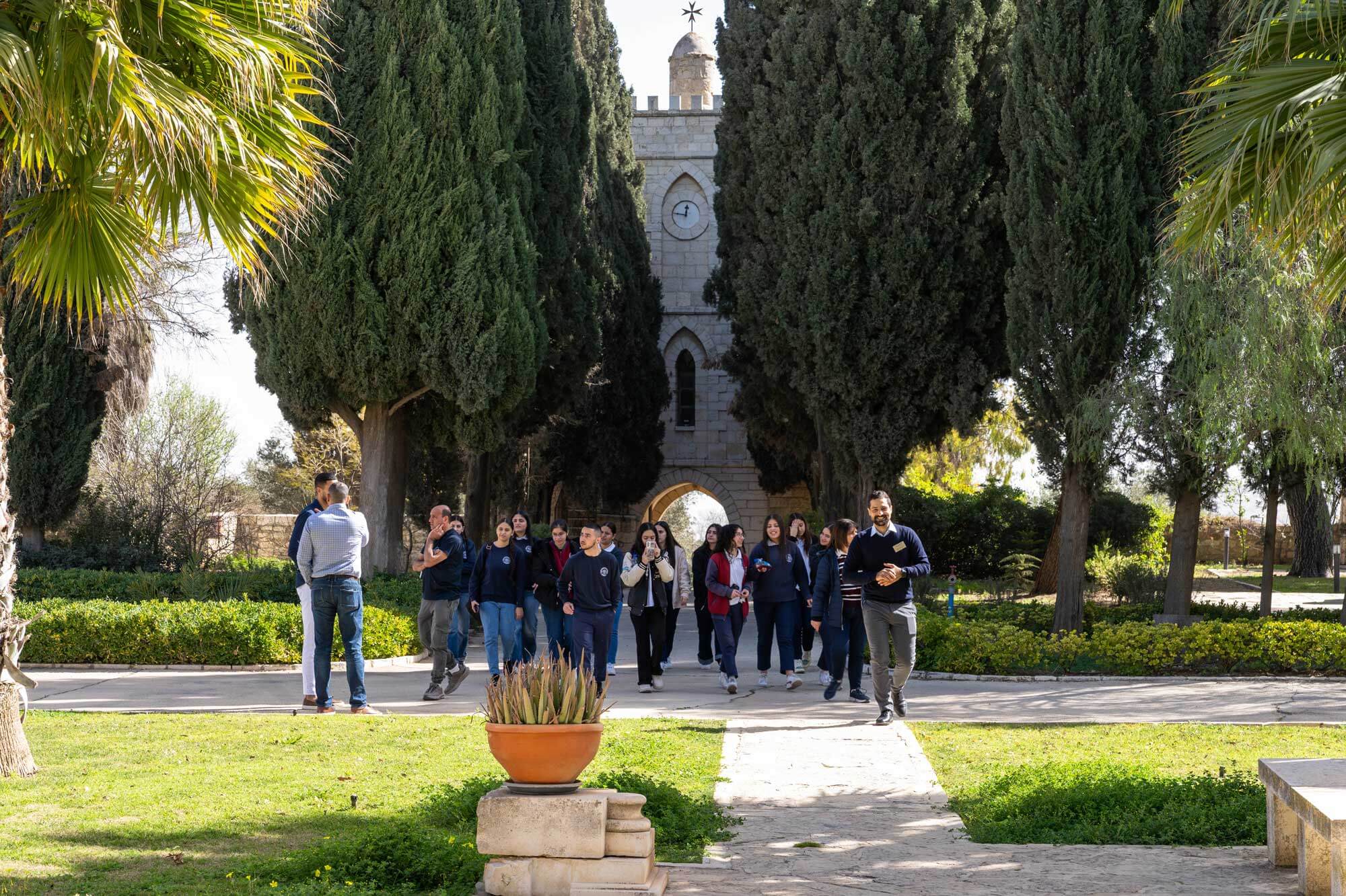 Students and a smiling guide walk toward a stone clock tower with an archway, framed by tall green cypress trees.