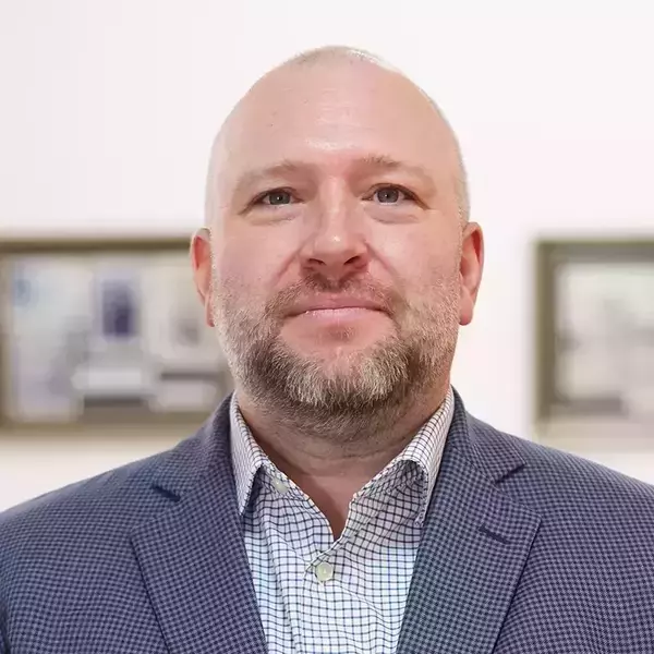 Gabriel Mitchell. Headshot of a bald man with a light beard, wearing a blue patterned blazer and a white and blue checkered shirt, looking forward.
