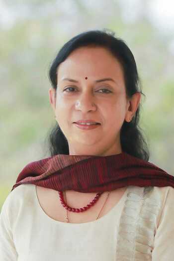 Smiling woman with dark hair and a red bindi wears a red and black striped shawl and a red beaded necklace.
