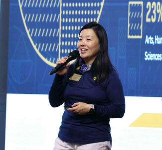 A smiling woman in a blue Notre Dame polo shirt with a gold shamrock logo speaks into a microphone at a presentation.