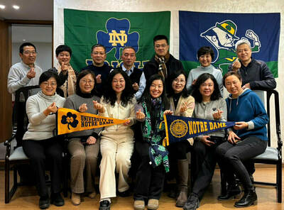 Fifteen smiling adults, some holding Notre Dame pennants, make heart gestures in front of ND and Fighting Irish flags.