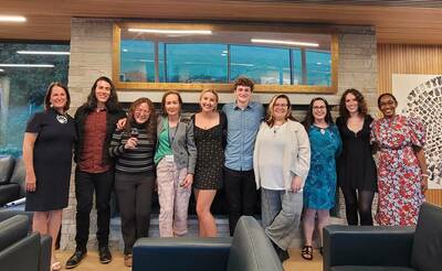 Ten smiling people, diverse in age and gender, stand together in a modern lounge with a stone wall and large window.