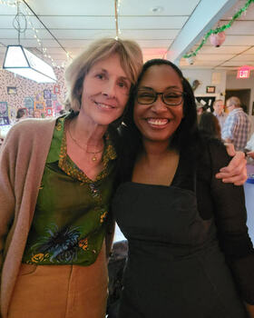Two smiling women, one blonde in a green patterned shirt, the other with dark hair and glasses, embracing indoors.