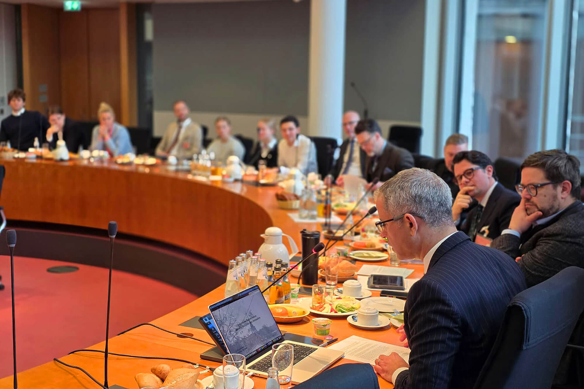Group of professionals attend a meeting at a curved orange wooden table, with laptops, microphones, papers, and refreshments.