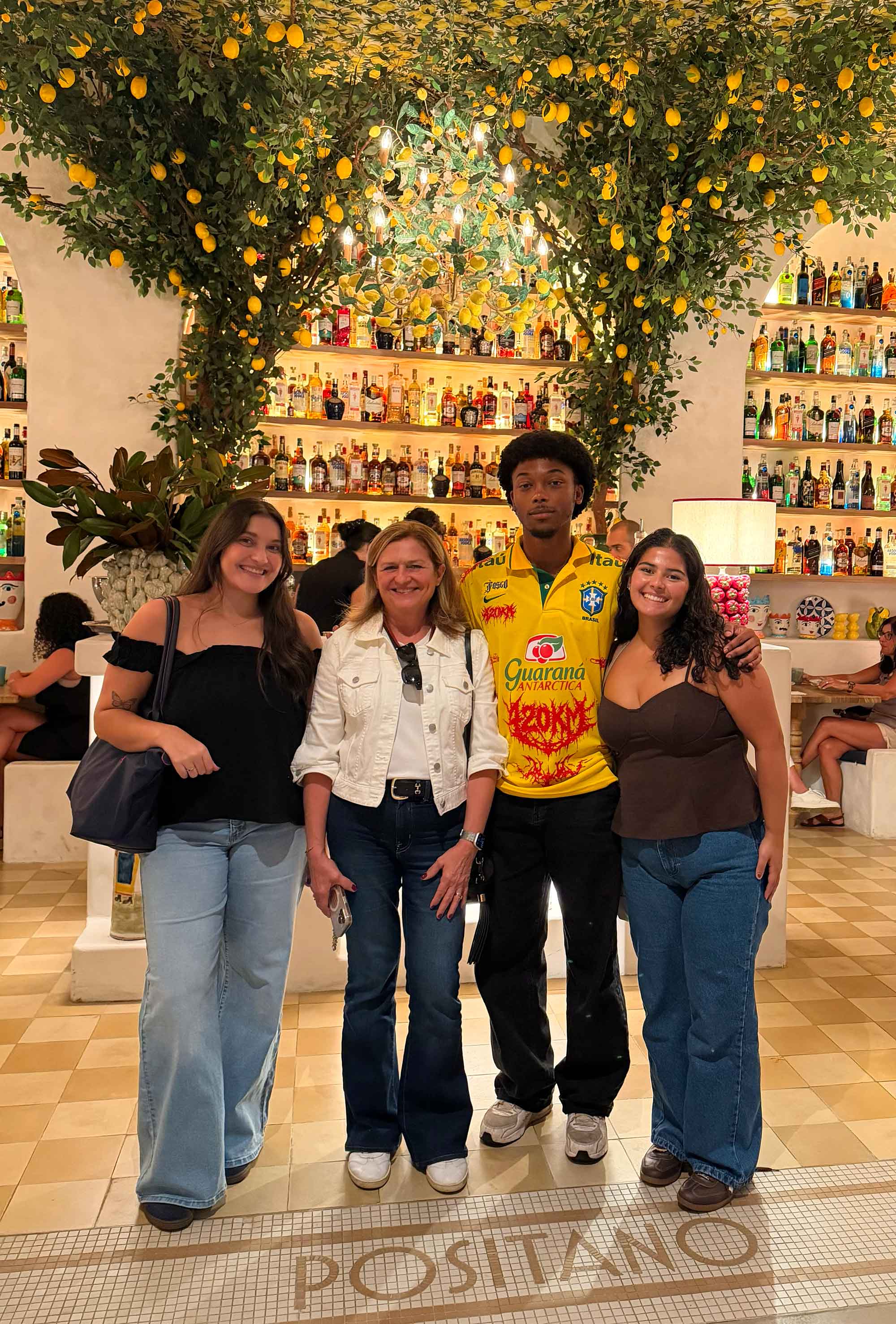 Four smiling people, including a man in a yellow Brazil jersey, stand in a Positano restaurant with lemon trees and liquor bottles.