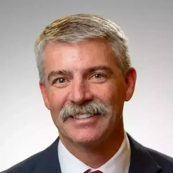 A smiling man with gray hair and a gray mustache wears a dark suit, white shirt, and red striped tie.