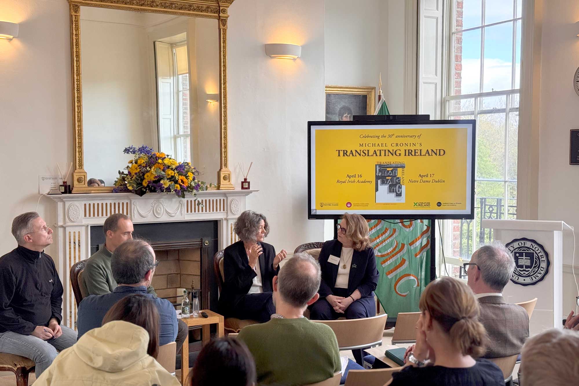 Two women speak at a panel, facing an audience. A screen behind them advertises "Translating Ireland" at Notre Dame Dublin.