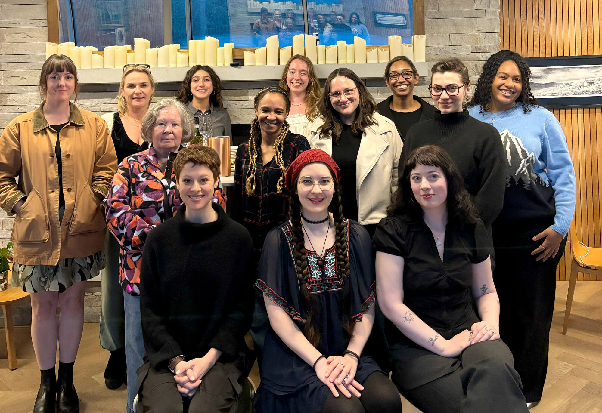 A diverse group of 14 smiling women stand and sit in a modern room with candles and a wooden wall.