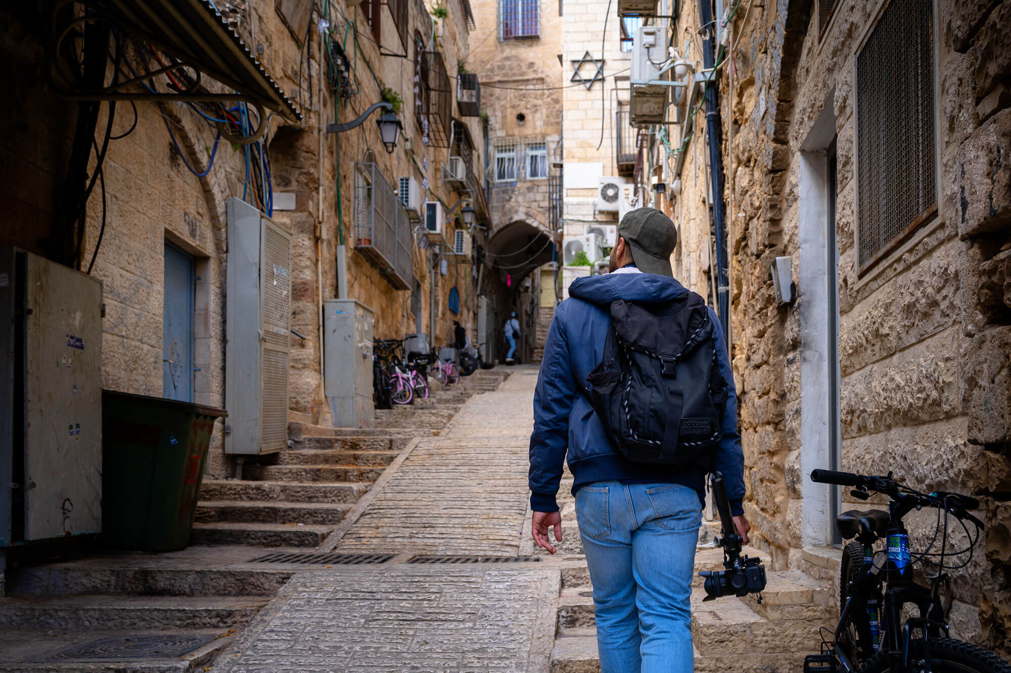 Person in blue jacket, jeans, and green cap with a backpack and camera walks up a narrow stone alleyway in Jerusalem.
