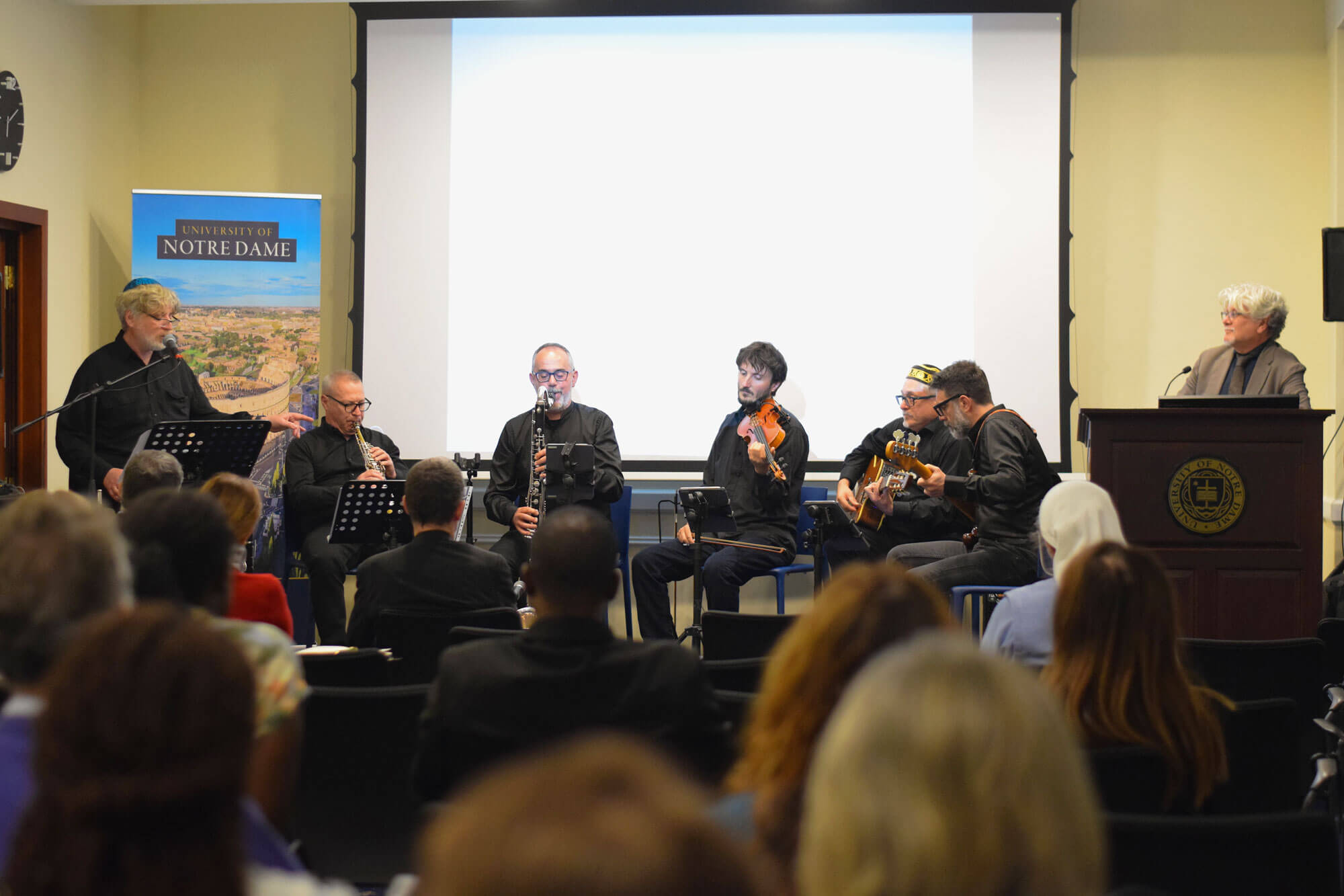 Five musicians play woodwinds, violin, and guitars for an audience at a University of Notre Dame event, with speakers on either side.