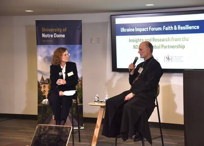 A woman with curly hair and glasses, and a bearded priest, both holding microphones, speak at the Notre Dame Ukraine Impact Forum.