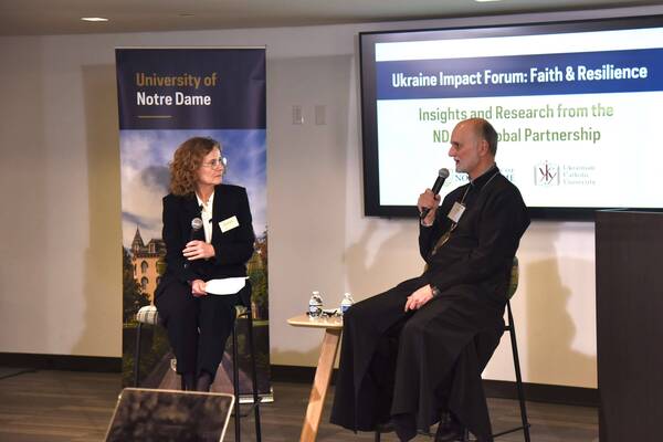 A woman with curly hair and glasses, and a bearded priest, both holding microphones, speak at the Notre Dame Ukraine Impact Forum.