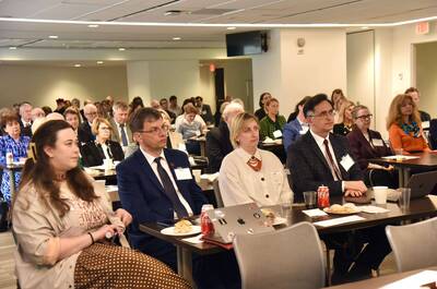 Audience sits attentively at tables with laptops in a brightly lit conference room.