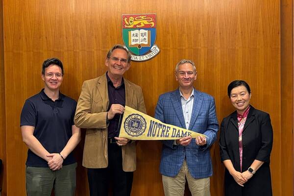 Four individuals smile, two men holding a cream and blue Notre Dame pennant, in front of the University of Hong Kong crest.